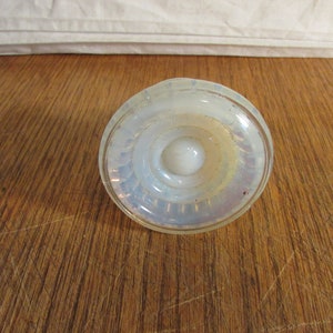May include: A clear, opalescent glass bottle stopper with a circular design. The stopper has concentric rings and a rounded top, creating a layered effect. The glass has a slight iridescence, reflecting light. The stopper is on a wooden surface.