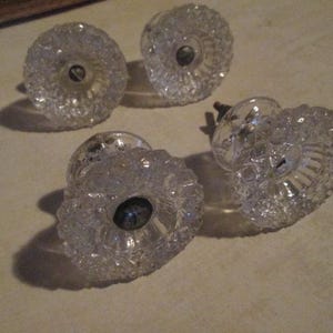 May include: Four clear glass cabinet knobs with a textured, faceted design. Each knob has a dark center and a metal screw for attachment. The knobs are likely for furniture restoration or decorative purposes.