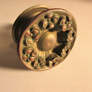 May include: Antique brass door knob with a floral design. The knob features a circular face with detailed relief carvings of stylized flowers and leaves. The central portion of the knob is slightly raised and smooth, while the edges show signs of age.