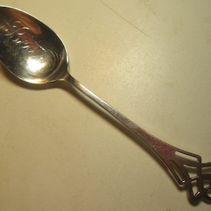 May include: A silver spoon with an ornate handle design. The spoon's bowl has an inscription. The handle features a decorative openwork pattern. The spoon is made of a shiny, reflective metal, likely stainless steel.