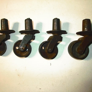 May include: Four antique metal caster wheels. Each wheel has a dark brown, cylindrical stem, a circular plate, and a small wheel. The wheels show signs of age and wear, with a rusty appearance.