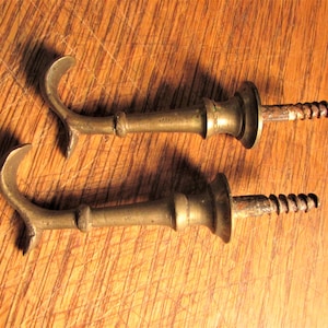 May include: Two antique brass hooks with a screw on the end. The hooks are in the shape of a curved 'S' and have a decorative finial at the top.
