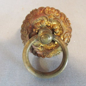 May include: Ornate, antique brass door knocker with a circular pull ring. The knocker features a detailed, golden lion's head design with intricate patterns. The ring is a muted brass color.