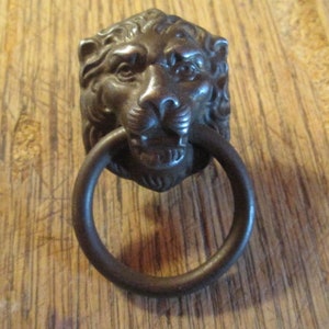 May include: A bronze-colored lion head door knocker with a circular handle. The lion's face is detailed with a mane, eyes, nose, and mouth. The ring handle is attached to the lion's mouth.