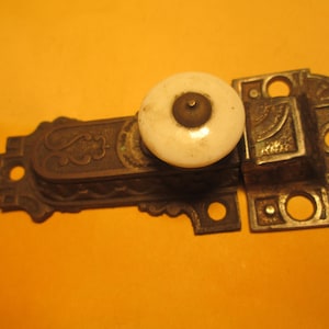 May include: An antique door latch with a white ceramic knob and ornate metalwork. The latch is composed of two parts, one with a decorative design and the other with a square shape. The metal has a tarnished, aged appearance.