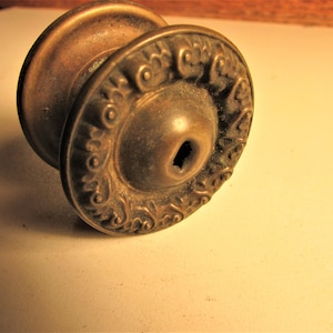 May include: Antique brass door knob with ornate floral detailing around the circular plate. The knob has a cylindrical base and a decorative, aged finish. A vintage hardware piece.