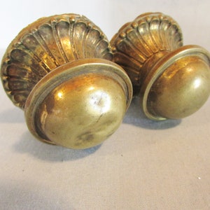 May include: Two antique brass-colored cabinet knobs. Each knob features a rounded base and a decorative, shell-like top. The knobs have a vintage aesthetic and are likely for furniture restoration or decorative purposes.