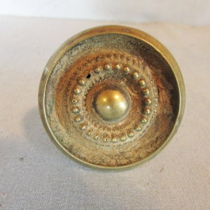 May include: A vintage brass knob with a circular design. The center features a raised, rounded element, surrounded by a ring of small, raised dots. The outer edge is a wider band with a slightly weathered appearance, suggesting age.