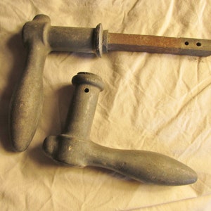 May include: Two antique-style door handles made of weathered gray metal. One handle features a long, rectangular iron spindle with two small circular holes at the end, while the other handle has a shorter, cylindrical neck with a single hole.