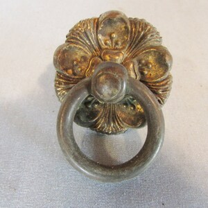 May include: Antique brass-toned drawer pull with a floral design and a circular handle. The hardware has a weathered appearance, suggesting age and vintage charm. Ideal for furniture restoration or adding a classic touch.