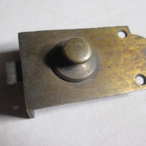 May include: Antique brass-toned cabinet latch with a rounded knob and rectangular base. The latch has a curved edge and two screw holes. The metal shows signs of age and wear, adding to its vintage appeal.
