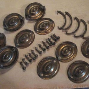 May include: A collection of antique brass hardware components, including oval drawer pulls, curved handles, and screws. The hardware has a tarnished, aged appearance, suggesting vintage or salvaged pieces. The components are arranged on a light-colored surface.