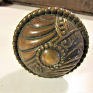 May include: A detailed, antique bronze door knob with an ornate, raised design. The knob features a central circular element, surrounded by intricate patterns and a beaded border. The color is a warm, aged bronze.