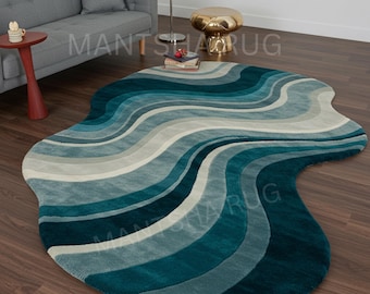 Puede incluir: Una alfombra de forma irregular con un degradado de rayas onduladas en tonos azul verdoso profundo, azul claro y crema. La alfombra tiene una textura suave y afelpada, siguiendo una silueta fluida y orgánica sobre el suelo.