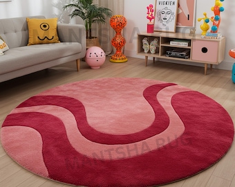 May include: A round rug with a wavy design in shades of pink and red. The rug features a soft, plush texture and is placed on a wooden floor. The design includes a light pink center, bordered by medium and dark red waves.
