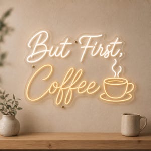 May include: A wall-mounted neon sign featuring the text But First, Coffee in a warm white script font. The sign includes a glowing outline of a coffee cup with steam rising from it, all mounted on a clear acrylic backing with visible hardware.