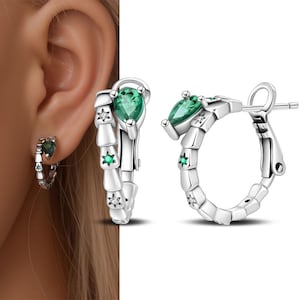 May include: Silver hoop earrings with a snake-like design. Each earring features a pear-shaped green gemstone at the top and a smaller green gemstone and a star-shaped accent. The earrings are shown on an ear and against a white background.