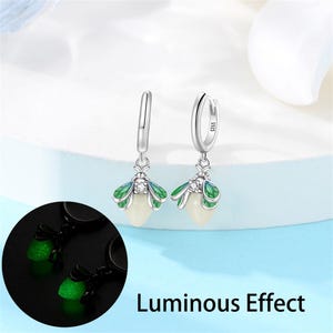 May include: Silver-tone hoop earrings with a firefly design. Each earring features a white pear-shaped body, green and blue wings, and a small clear gem. The image includes the text "Luminous Effect".