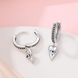 May include: A pair of silver hoop earrings with a dangling charm. Each charm is a heart-shaped design with a blue eye detail. The earrings have a dotted pattern along the edges.