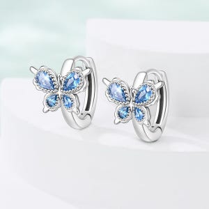 May include: A pair of silver-colored hoop earrings, each featuring a butterfly design. The butterflies have light blue, teardrop-shaped gemstones and are accented with small, round details. The earrings are displayed on a white surface.