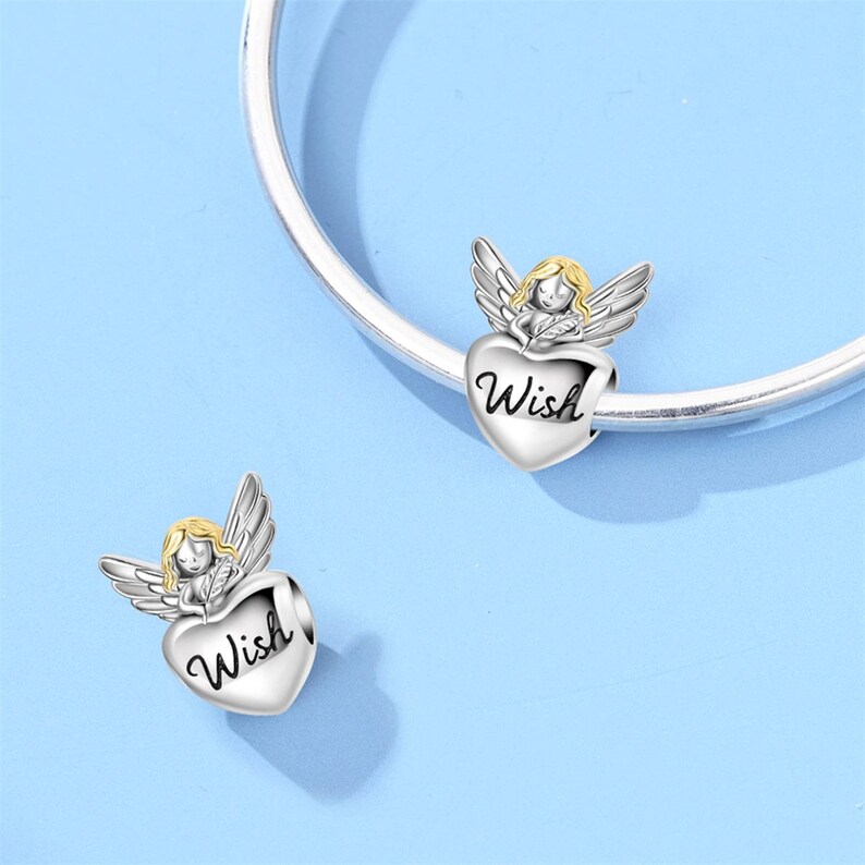May include: Two silver-toned angel charms with gold hair and wings, each holding a heart-shaped charm with the word "Wish" in cursive. The charms are displayed on a silver bracelet against a light blue background.