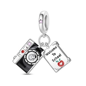 May include: A silver charm with a camera and a notepad. The black camera has a clear gem in the center and a pink gem on top. The notepad says "Welcome To School" with a red apple illustration.