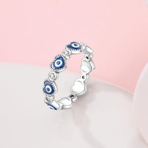 May include: Silver ring featuring a repeating pattern of heart-shaped charms and blue evil eye charms. The ring has a polished finish and is set against a soft pink and white background. The evil eye charms have a blue iris and a white pupil.