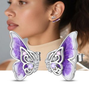 May include: Silver butterfly earrings with purple and white enamel wings. The earrings feature clear and pink gemstones. One earring is worn on the ear, and the other is displayed in front of the ear.
