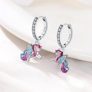 May include: A pair of silver hoop earrings with a row of small clear stones. Each earring has a dangling, colorful unicorn charm with pink, blue, and purple accents. The earrings are on a white surface.