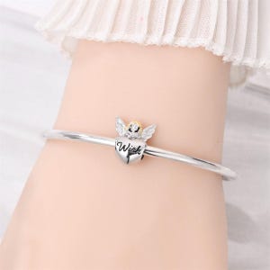 May include: A silver bangle bracelet with a heart-shaped charm. The charm features an angel with wings and the word "Wish" inscribed on the heart. The angel has a gold-colored head and the wings are silver.