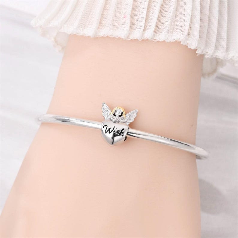 May include: A silver bangle bracelet with a heart-shaped charm. The charm features an angel with wings and the word "Wish" inscribed on the heart. The angel has a gold-colored head and the wings are silver.