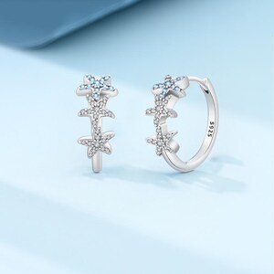 May include: A pair of silver hoop earrings featuring three starfish motifs along the front. Each starfish is encrusted with small clear stones, and the top starfish on each hoop is accented with light blue stones. The inner band is stamped with S925.