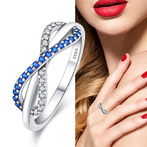 May include: A silver-colored ring with an infinity design. The ring features a band of blue gemstones and a band of clear gemstones. The ring is marked with "S925".
