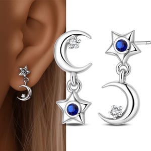 May include: Silver-toned dangle earrings featuring a crescent moon and star design. The star has a blue gemstone center, and the moon has a small, clear gemstone. The earrings are shown on an ear and also displayed separately.
