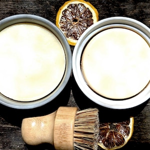 Puede incluir: Dos jabones redondos blanquecinos en cuencos grises y blancos, con un cepillo de madera y rodajas de limón secas. Los jabones tienen una superficie lisa. El cepillo tiene un mango de bambú y cerdas naturales. La disposición está sobre una superficie de madera oscura.