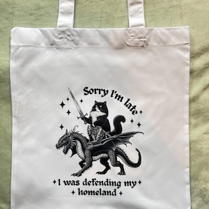 May include: White tote bag with a black and white graphic of a cat in armor riding a dragon, holding a sword. Text reads "Sorry I'm late + I was defending my + homeland +."