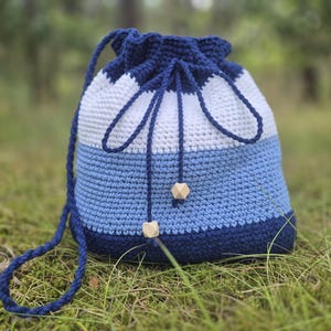 Può includere: Una borsa a coulisse all'uncinetto fatta a mano nei toni del blu e del bianco. La borsa presenta una parte superiore e inferiore blu navy, con una striscia bianca al centro. Il cordoncino è blu navy e la borsa ha accenti di perline di legno. La borsa è sull'erba verde.