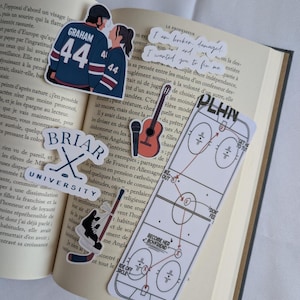 Op de afbeelding: Een verzameling stickers op een open boek. Stickers bevatten een stel in hockeytruien, een gitaar, een hockeybaan diagram en tekst die luidt "I am broken, damaged. I wanted you to fix me."