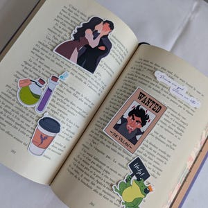 Peut inclure: Livre ouvert avec plusieurs autocollants colorés servant de marque-pages. Les autocollants incluent un couple s'embrassant, une affiche de recherche avec un méchant, une grenouille avec un panneau "Help!" et des motifs scientifiques. Les pages du livre sont remplies de texte français.