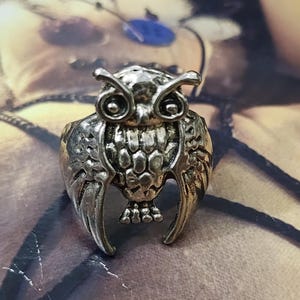 May include: A silver-toned metal ring featuring a detailed owl design. The owl has prominent eyes, textured feathers on its chest, and wings that extend downward along the sides of the band. The metal has an antiqued finish with dark accents.