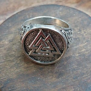 Könnte beinhalten: Ein silberfarbener Metallring mit einer kreisförmigen Vorderseite, auf der ein erhabenes Valknut-Symbol vor einem texturierten, dunklen Hintergrund zu sehen ist. Die Ringschiene zeigt komplexe, geprägte Knotenmuster an den Seiten.