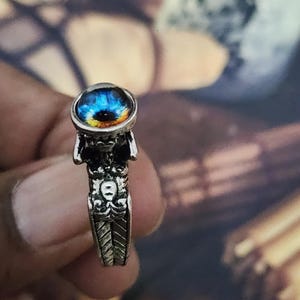 Op de afbeelding: Een zilverkleurige metalen ring met een opvallende ronde cabochon met een realistisch blauw en oranje irisontwerp. De ringband is gedetailleerd met een reliëf van een skeletfiguur en ingewikkelde patronen die langs de zijkanten doorlopen.
