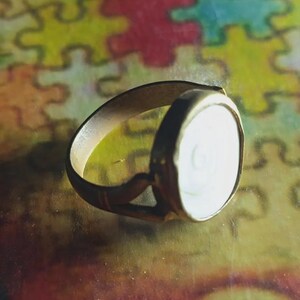 May include: A gold-toned metal ring featuring a circular, white, flat inlay with a subtle, centered spiral pattern. The band of the ring has a split design near the setting, and the metal surface shows a warm, slightly burnished finish throughout.