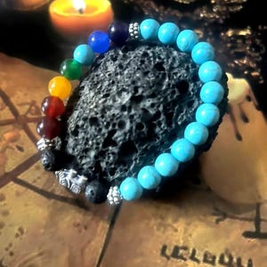 May include: A beaded bracelet featuring turquoise and multicolored beads, displayed on a textured black stone. The bracelet has silver-colored accents and is set against a backdrop with a lit candle.
