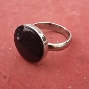 May include: A silver ring with a large, oval, black stone. The ring band is smooth and shiny, and the stone is set in a simple bezel. The ring is displayed on a red surface.