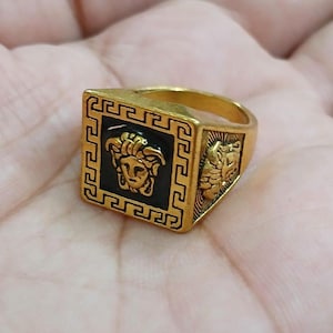 Puede incluir: Anillo cuadrado dorado con un diseño de cabeza de Medusa sobre un fondo negro. El anillo tiene un patrón geométrico alrededor de la cabeza de Medusa y en los lados. El anillo se sostiene en una mano.