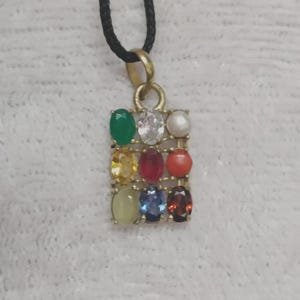 Op de afbeelding: Een rechthoekige goudkleurige hanger aan een zwart koord, met een raster van negen ovale edelstenen in verschillende kleuren, waaronder groen, transparant, wit, geel, rood, oranje, lichtgeel, blauw en donkerrood, gerangschikt in drie rijen.