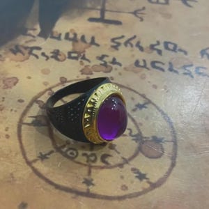 May include: A black ring with a gold-colored bezel and a large, oval, purple gemstone. The ring sits on a surface with faded text and symbols.