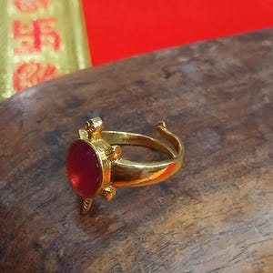 以下が含まれることがあります： 亀の形をしたデザインのゴールドトーンの金属製リング。甲羅の中央には滑らかで丸い濃い赤色の石がはめ込まれています。リングのバンドは後ろが開いており、サイズ調整が可能で、金属部分は光沢のある磨き上げられた仕上げになっています。