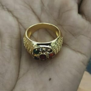 Op de afbeelding: Een gouden ring met een decoratieve band en een centrale cluster van kleurrijke edelstenen. De band van de ring heeft een textuurontwerp. De edelstenen omvatten rood, groen en andere kleuren. De ring wordt op een hand getoond.